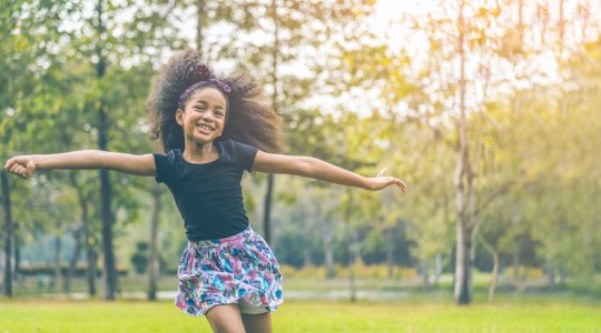 A little girl runs smiling with her arms out in a grassy field. 